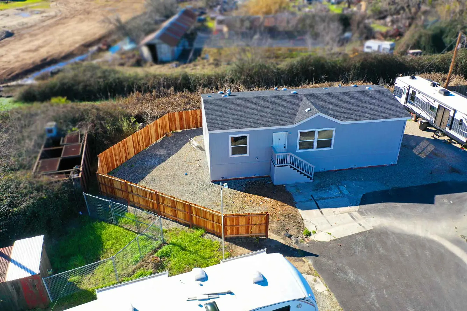 Aerial view of a light gray manufactured home with white entry steps and a fenced gravel yard, with travel trailers parked nearby.